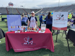 Person standing behind a table with nutrition information. The table is set up on a football field in a stadium