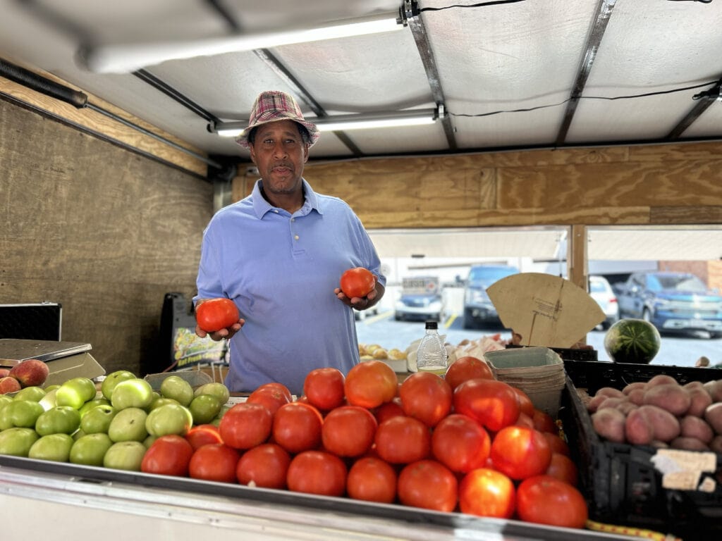 Man holding tomatoes at the outdoor farmer's market at UAMS. He is standing behind a table full of green and red tomatoes, potatoes, and other items.