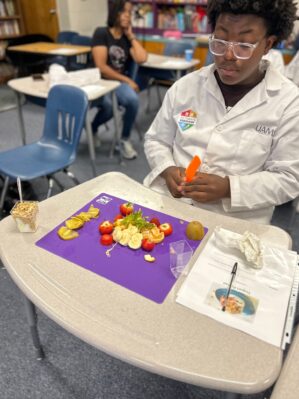 Pathways student, wearing a white UAMS coat, sits in front of a mat on a desk with a variety of cut up fruit